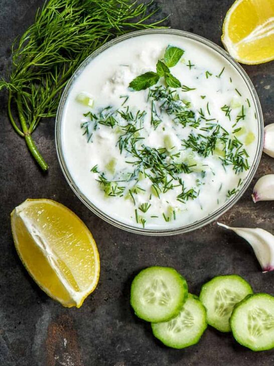a bowl of tzatziki sauce with fresh lemon, fresh cucumbers, garlic cloves, and fresh dill and mint around the bowl on a table