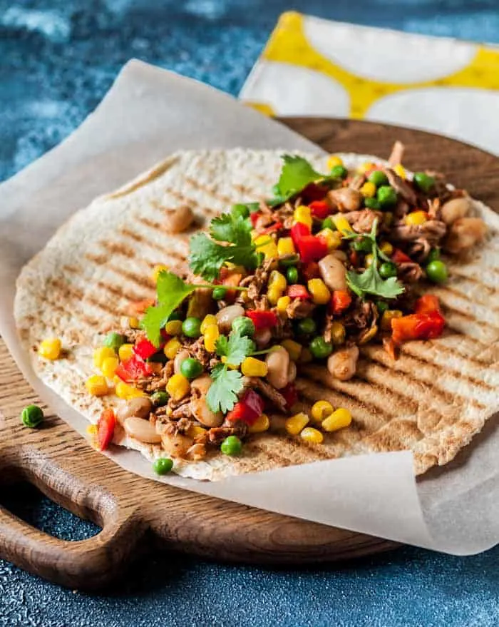 Pulled pork and grilled, chopped vegetables on a tortilla that's set on a round wooden board 