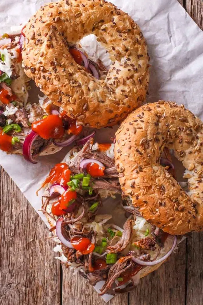 Two sliced bagels on parchment paper set on a wood table, topped with instant pot pulled pork and chopped red onion and bell pepper