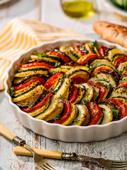 A vegetable tian just after baking in a round white dish on a white table with a striped yellow and white napkin