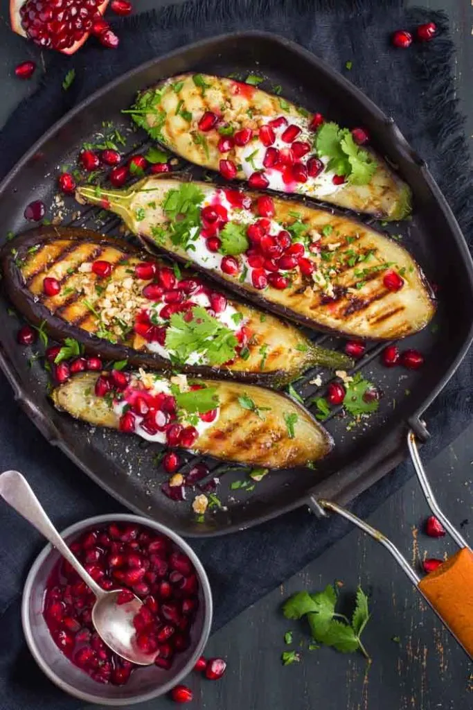 grilled eggplant topped with garlic yogurt sauce, walnuts and pomegranate with a small dish of pomegranate arils and a spoon next to it