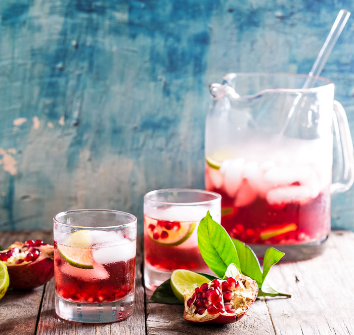 A pitcher of pomegranate margaritas with two glasses on a wooden board with a blue background