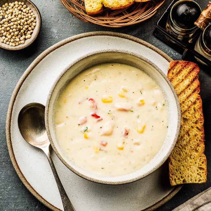 A bowl of seafood chowder with two toasted slices of french bread and a small dish of whole white pepper