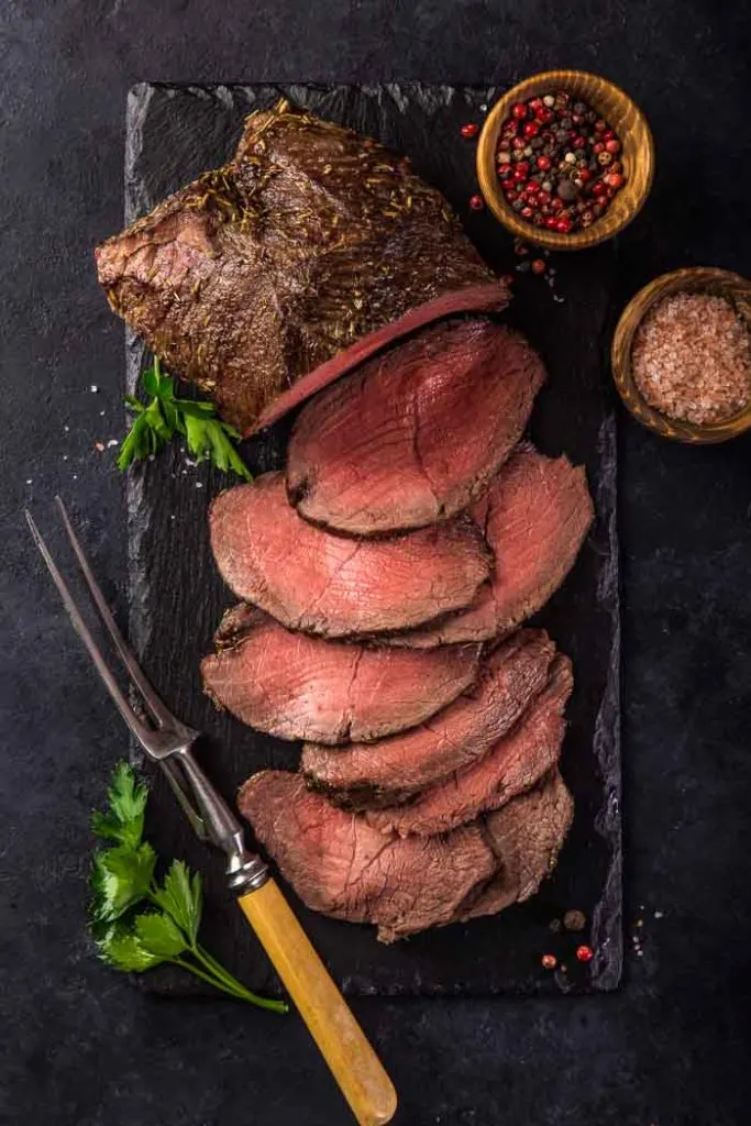 a beef roast with sliced roast beef next to it on a black plank with a small dish of each salt and peppercorns
