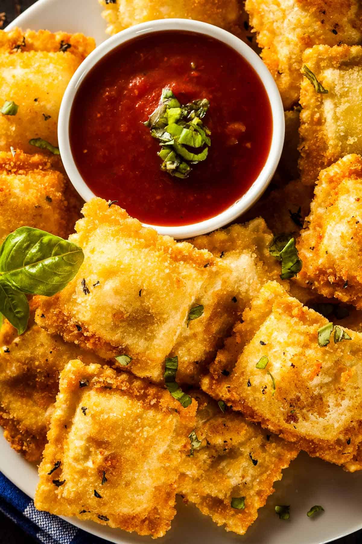 a white bowl filled with fried ravioli and a small white dish of marinara sauce with a sprig of fresh basil