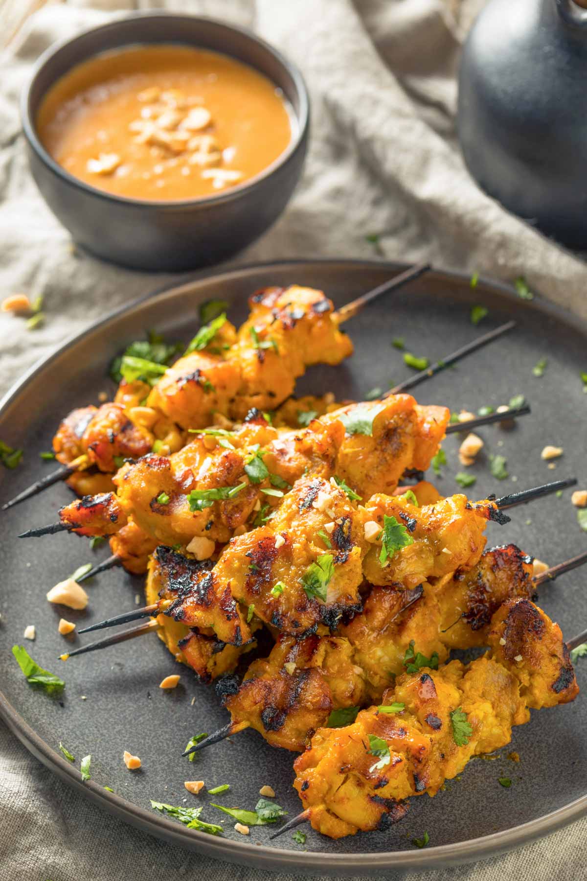 Chicken Satay Skewers + Peanut Sauce (Freezer-Friendly) an overhead shot of a black plate with a pile of chicken satay skewers on a tan linen napkin and a dish of peanut sauce