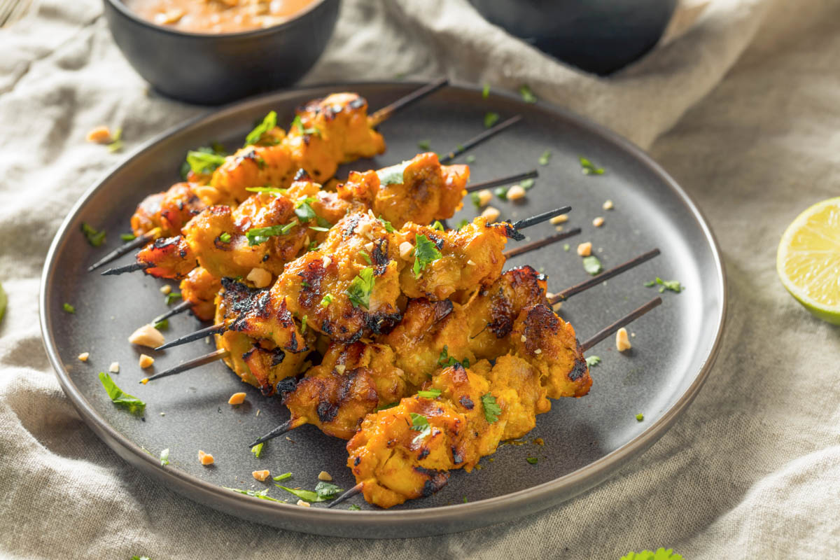 Chicken Satay Skewers + Peanut Sauce (Freezer-Friendly) a black plate with a pile of chicken satay skewers on a tan linen napkin