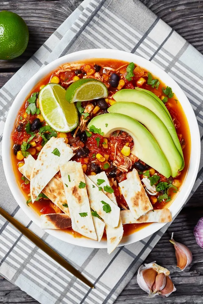 a closeup of a bowl of easy taco soup topped with homemade tortilla chips, avocado slices, and fresh lime wedges
