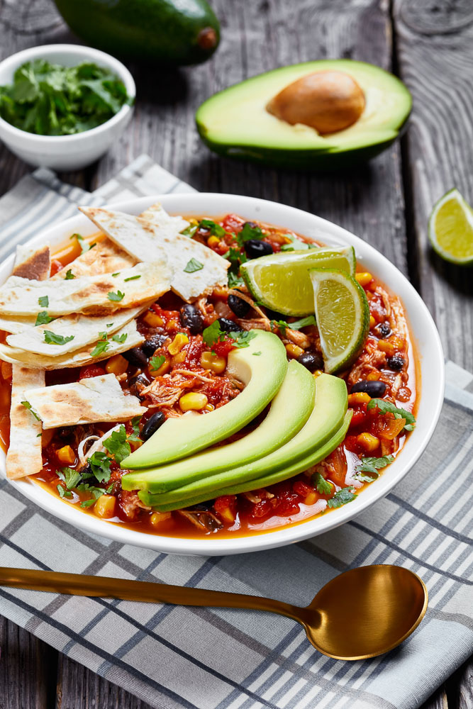 a bowl of easy taco soup topped with homemade tortilla chips, avocado slices, and fresh lime wedges, next to a halved avocado and a small dish of chopped cilantro
