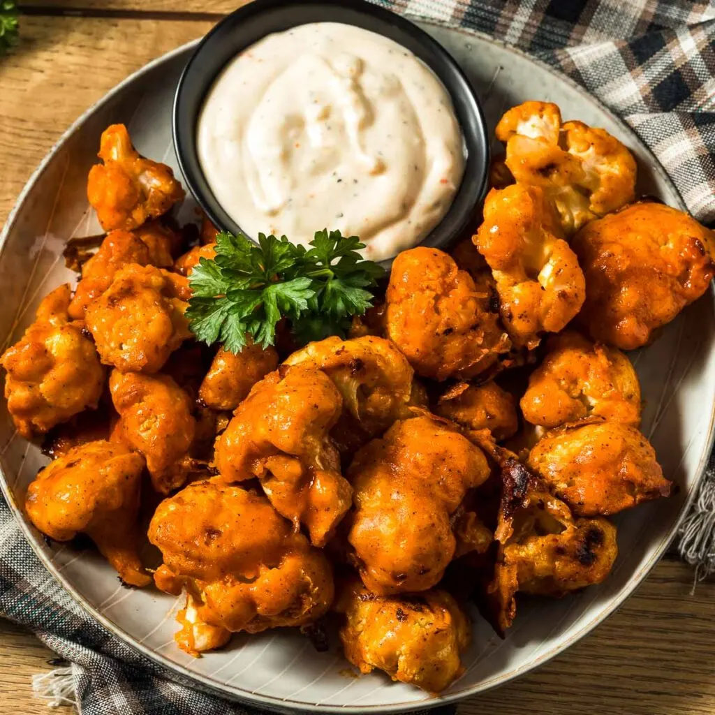 a plate of buffalo cauliflower wings with a small dish of ranch dressing next to a plaid towel