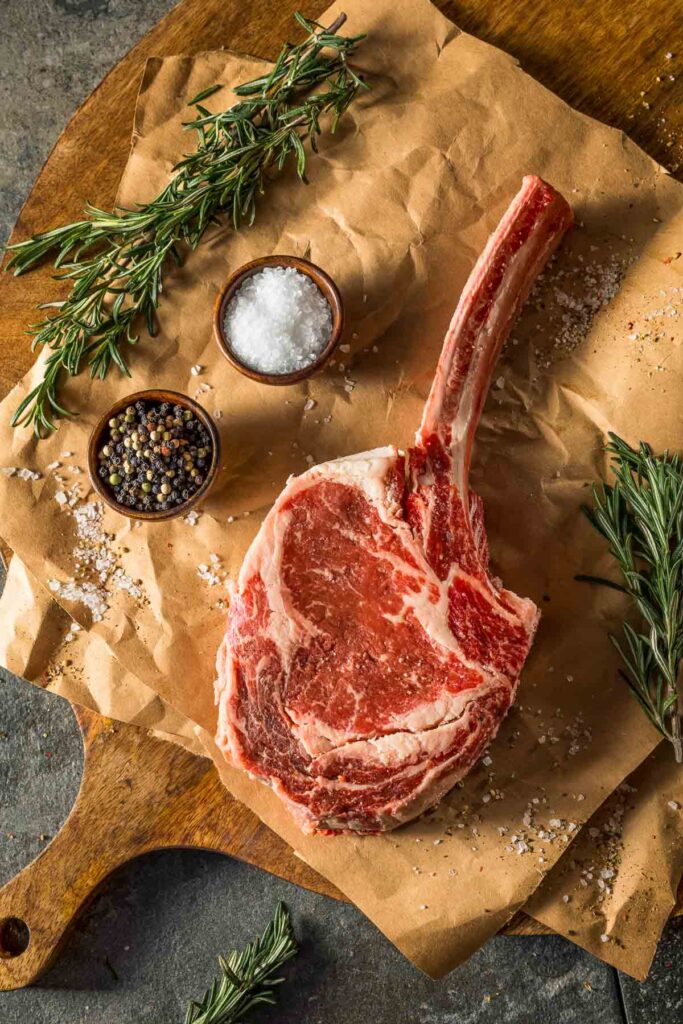 a tomahawk steak on parchment paper next to a dish of salt and pepper with fresh rosemary