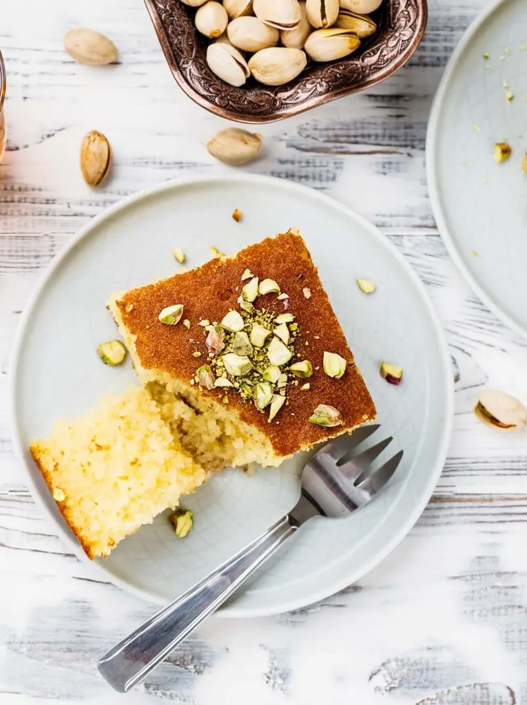 an overhead shot of a slice of revani turkish cake on a white plate garnished with pistachios
