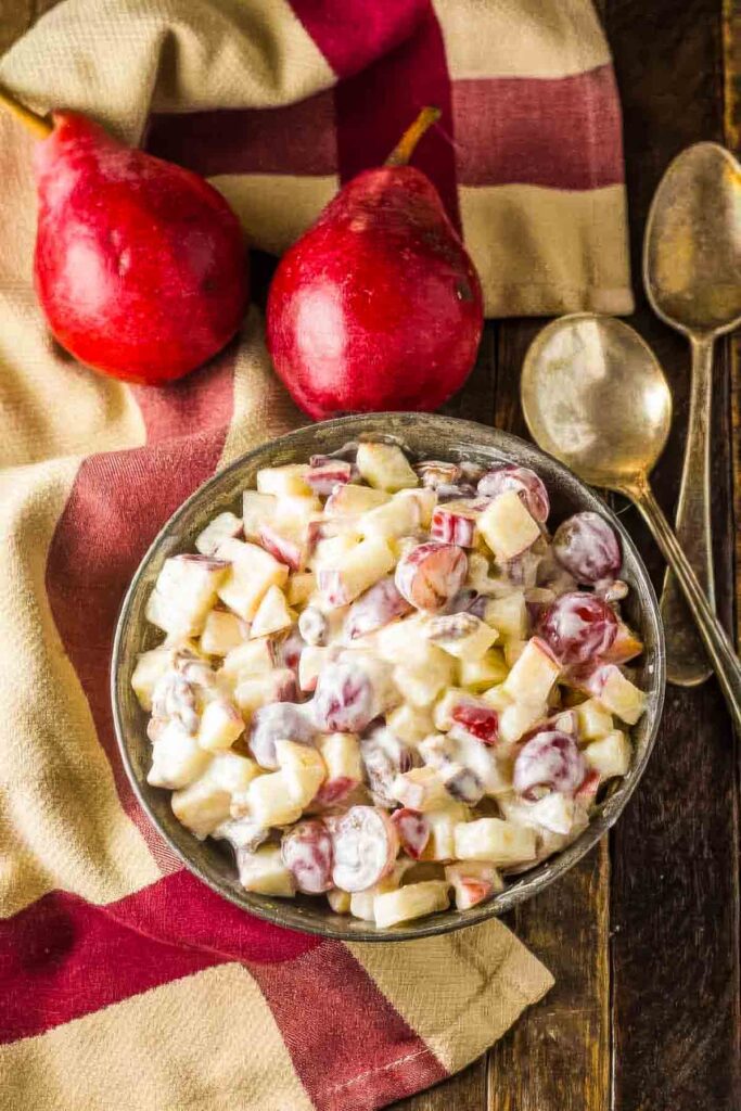 a metal bowl filled with fall fruit salad on a wooden table, next to a tan and burgundy towel, two silver spoons, and two red pears
