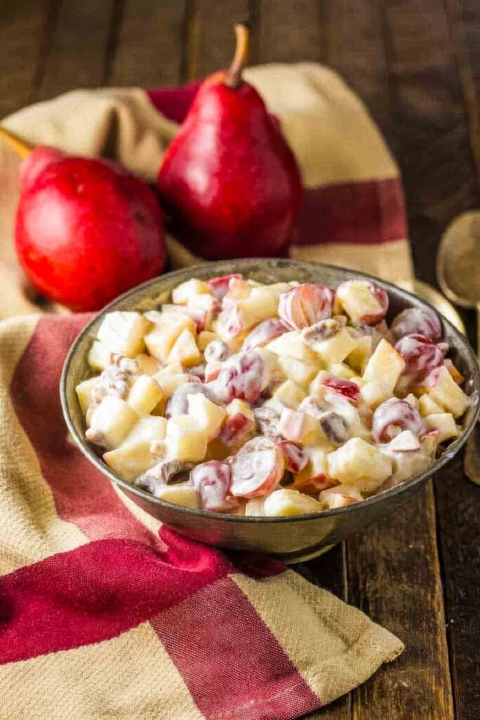 a metal bowl filled with fall fruit salad on a wooden table, next to a tan and burgundy towel and two red pears