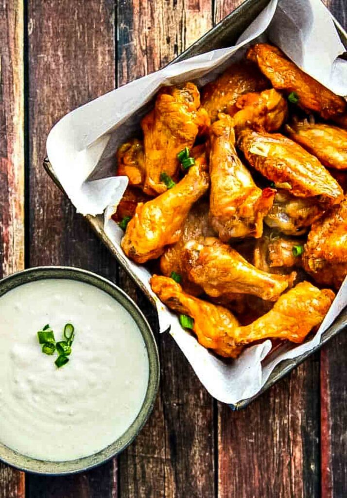 Crispy Baked Buffalo Wings buffalo wings just out of the oven are in a vintage metal dish lined with parchment paper next to a small dish of blue cheese dressing
