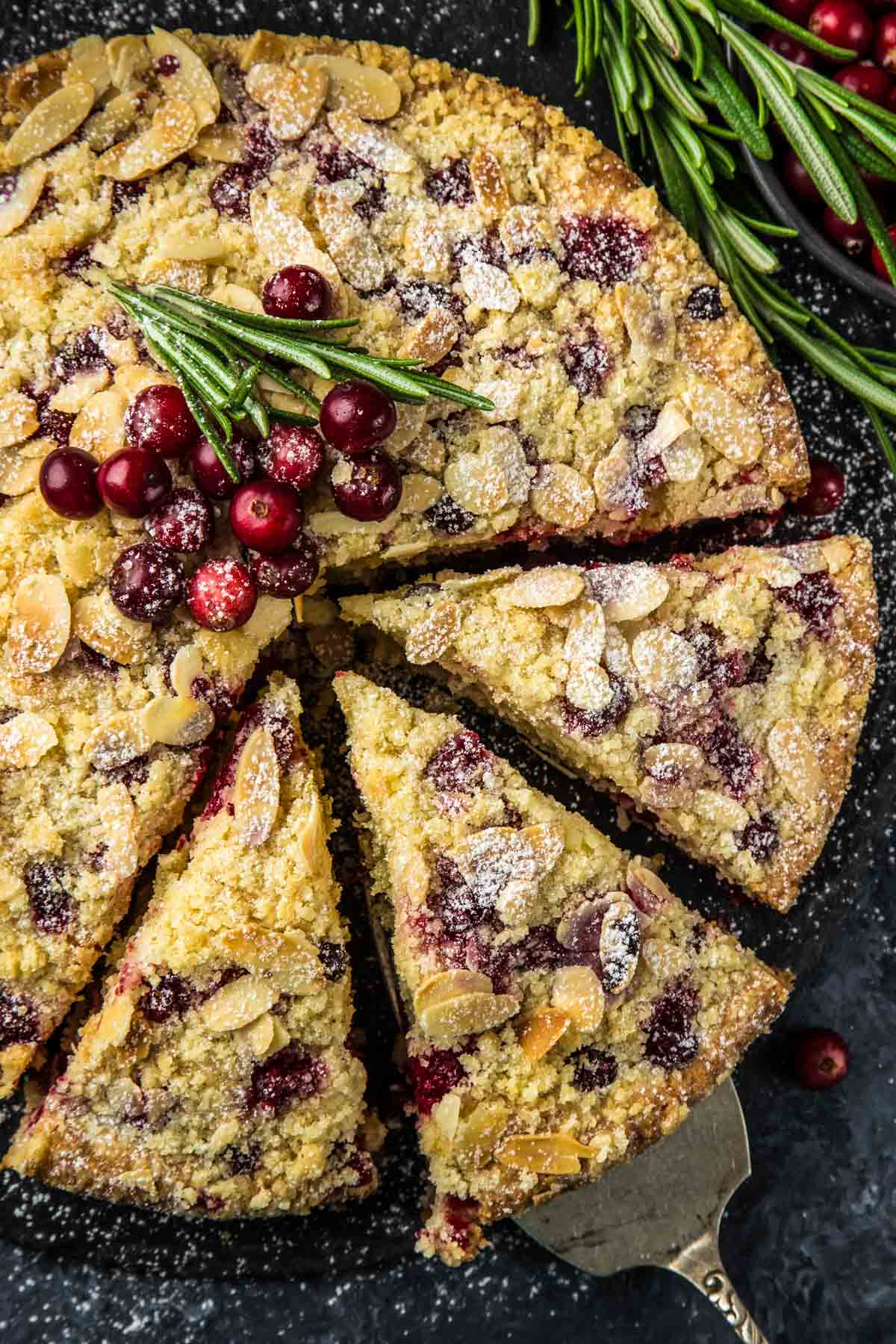 The Best Cranberry Christmas Cake (Make-Ahead Friendly) a closeup, top-down shot of a cranberry christmas cake topped with fresh cranberries and a sprig of rosemary with three slices cut and one slice on a vintage spatula