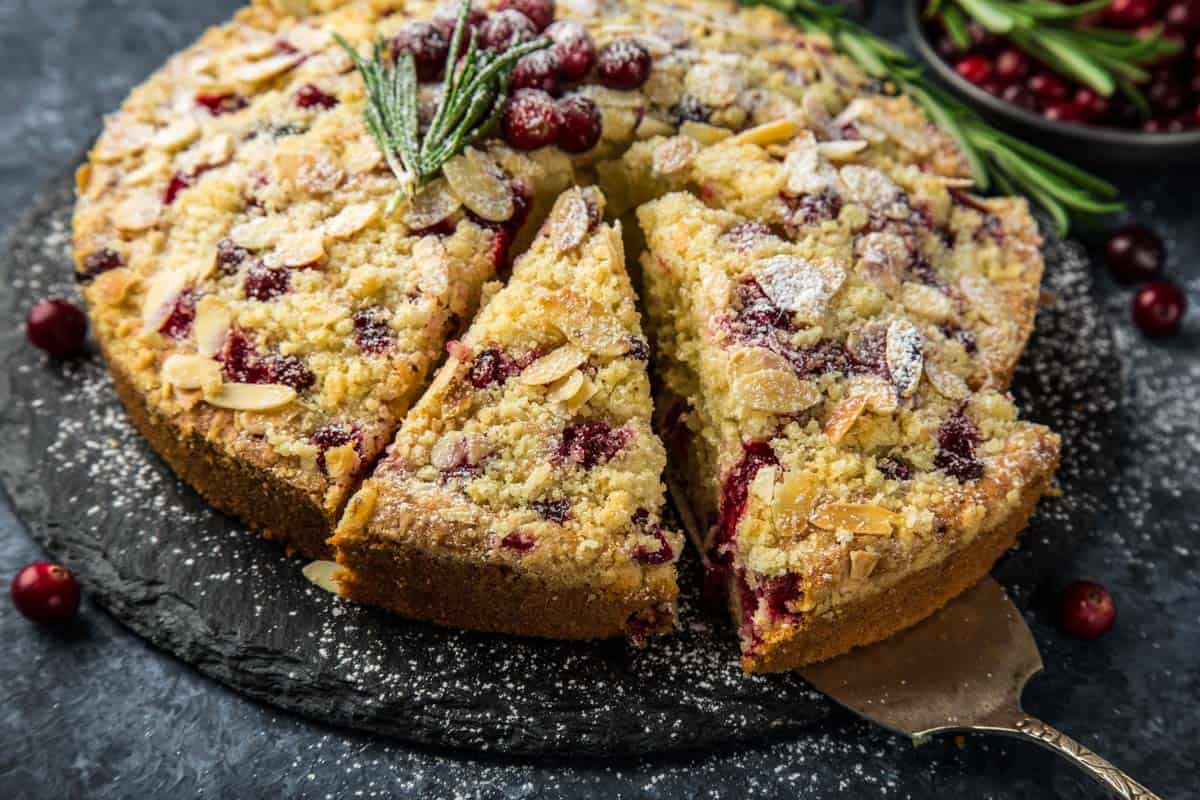 a side view of a cranberry christmas cake topped with fresh cranberries and a sprig of rosemary with three slices cut and one slice on a vintage spatula