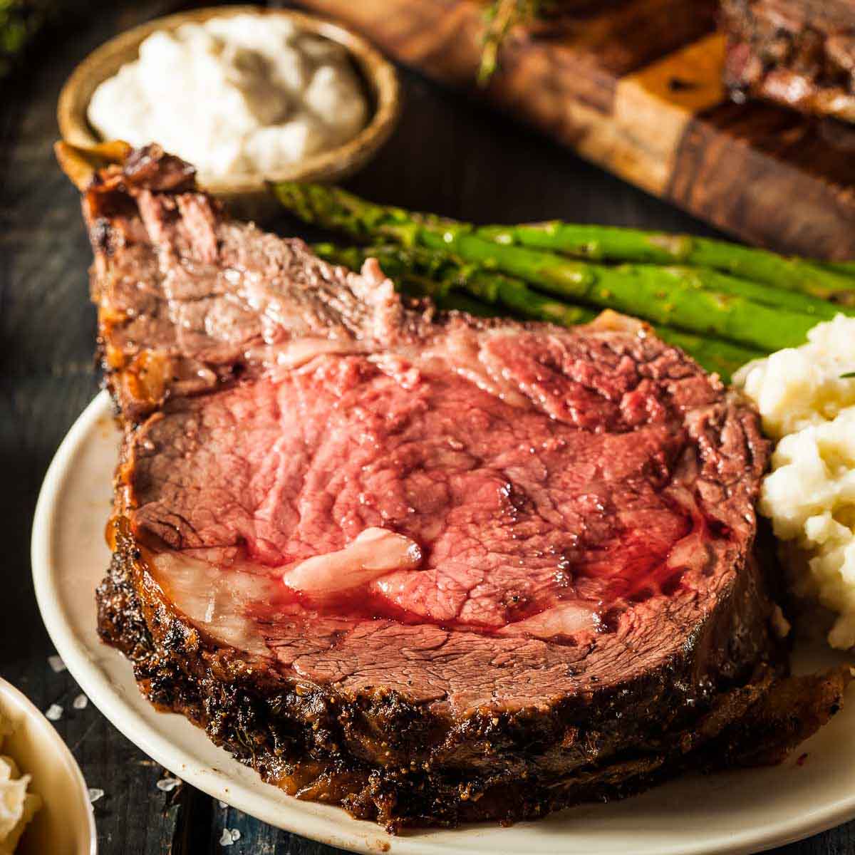 a slice of prime rib on a white plate with mashed potatoes and grilled asparagus, next to a small dish of horseradish sauce. the rest of the standing rib roast is in the background