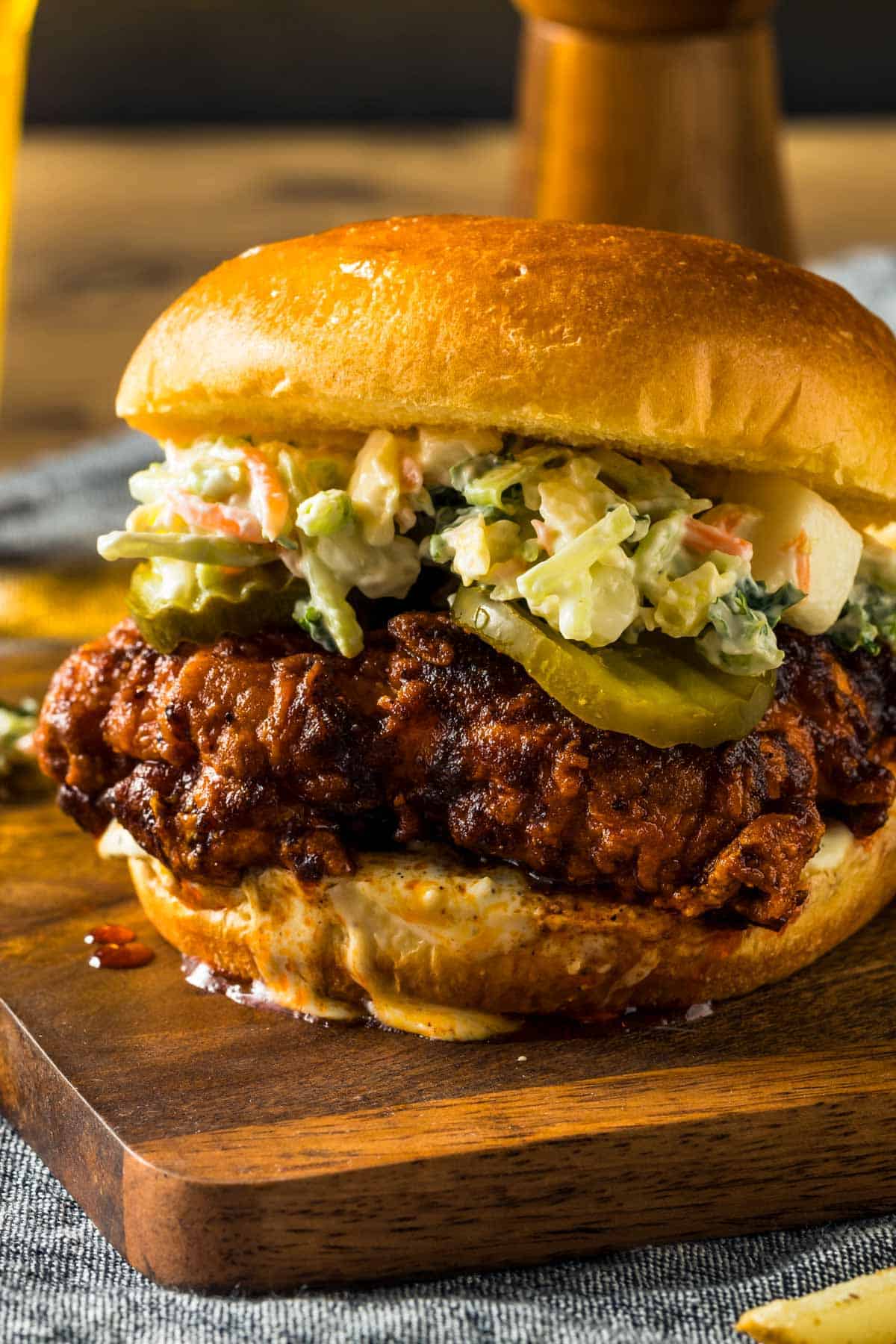 Nashville Hot Chicken Sandwich You Can Easily Make at Home a closeup of a nashville hot chicken sandwich topped with pickles and coleslaw on a wood cutting board
