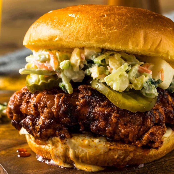 an extreme closeup of a nashville hot chicken sandwich topped with pickles and coleslaw on a cutting board