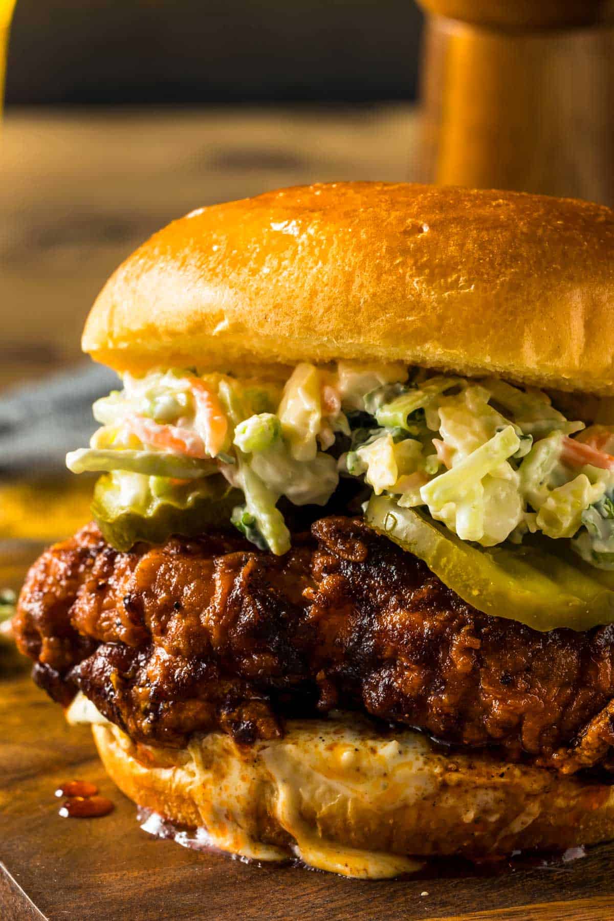 Nashville Hot Chicken Sandwich You Can Easily Make at Home a closeup of a nashville hot chicken sandwich dripping with sauce and topped with pickles and coleslaw on a cutting board