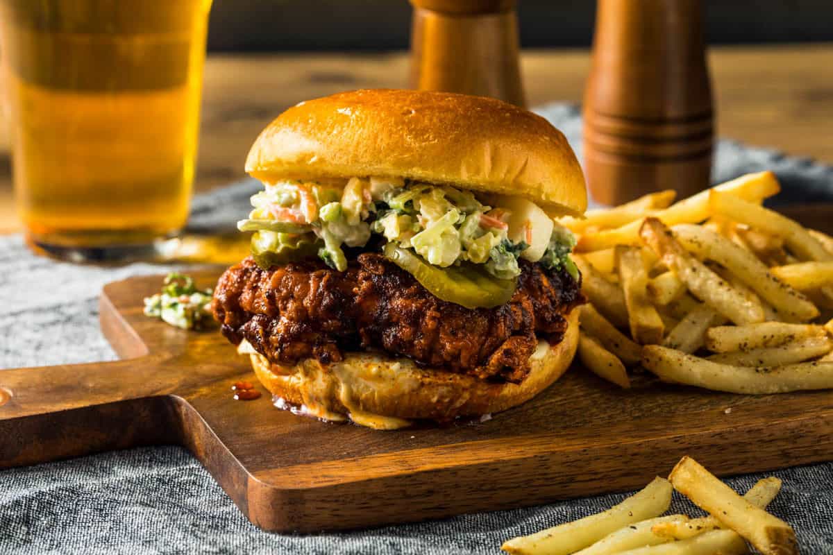 Nashville Hot Chicken Sandwich You Can Easily Make at Home a nashville hot chicken sandwich on a cutting board with french fries next to a cold glass of beer