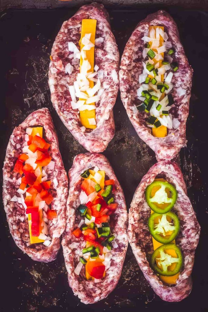 Raw bratwursts sliced open and stuffed with cheddar cheese, chopped onions, bell peppers, jalapeños, and other colorful vegetables, arranged on a dark baking sheet before being wrapped in bacon and cooked.