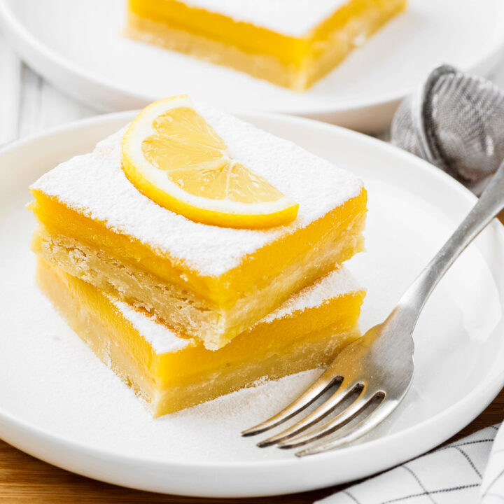 two lemon bars on a white plate with a fork, topped with powdered sugar and a fresh slice of lemon