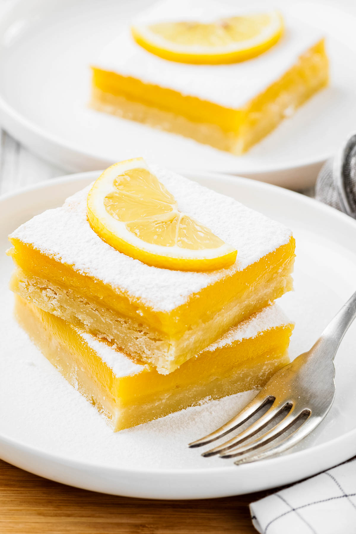 The Best Lemon Bars two lemon bars on a white plate with a fork, topped with powdered sugar and a fresh slice of lemon with another lemon bar on a plate in the background