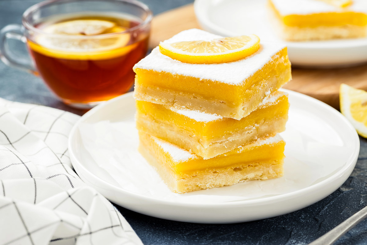 three lemon bars on a white plate, topped with powdered sugar and a fresh slice of lemon next to a cup of hot tea