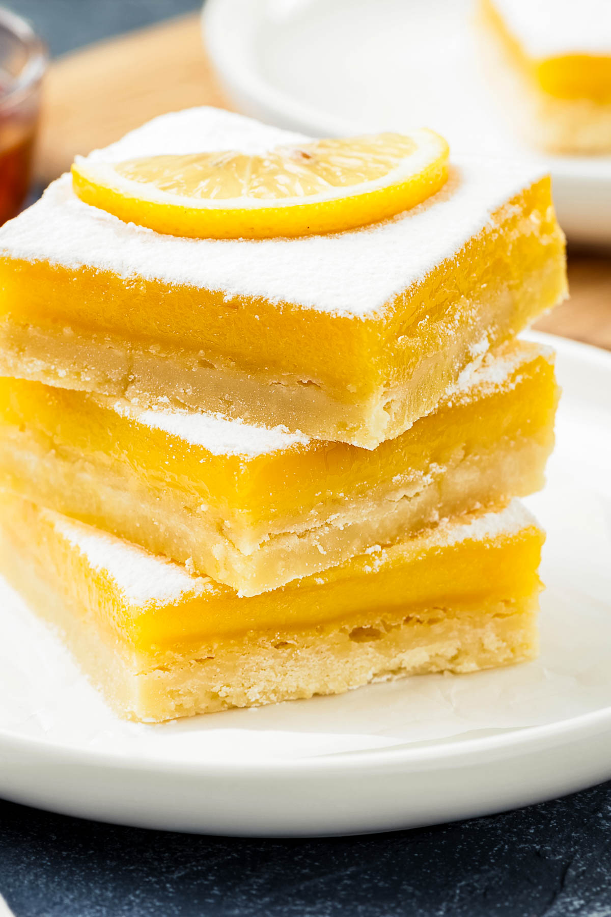three lemon bars stacked on a white plate, topped with powdered sugar and a fresh slice of lemon
