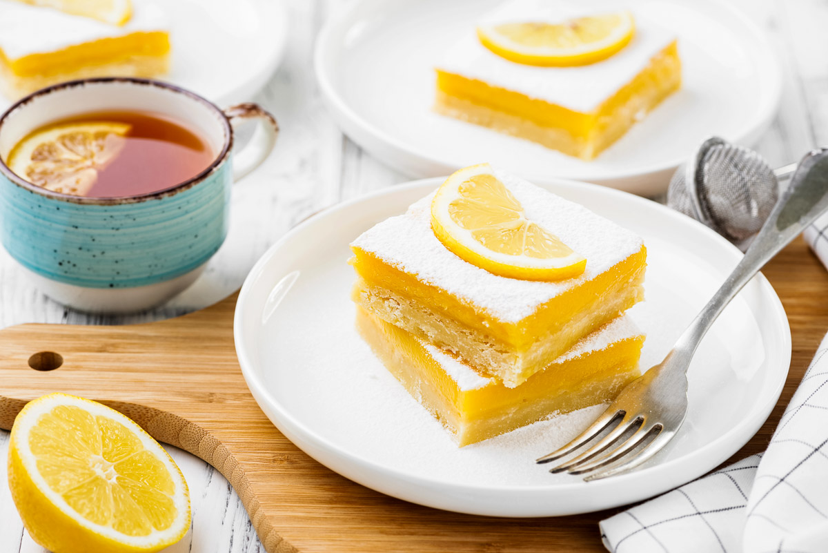 two lemon bars on a white plate with a fork, topped with powdered sugar and a fresh slice of lemon next to a cup of tea