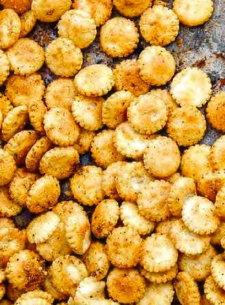 Close-up shot of crispy baked oyster crackers coated in Old Bay seasoning, showing golden edges and coarse spice texture on a dark baking tray.