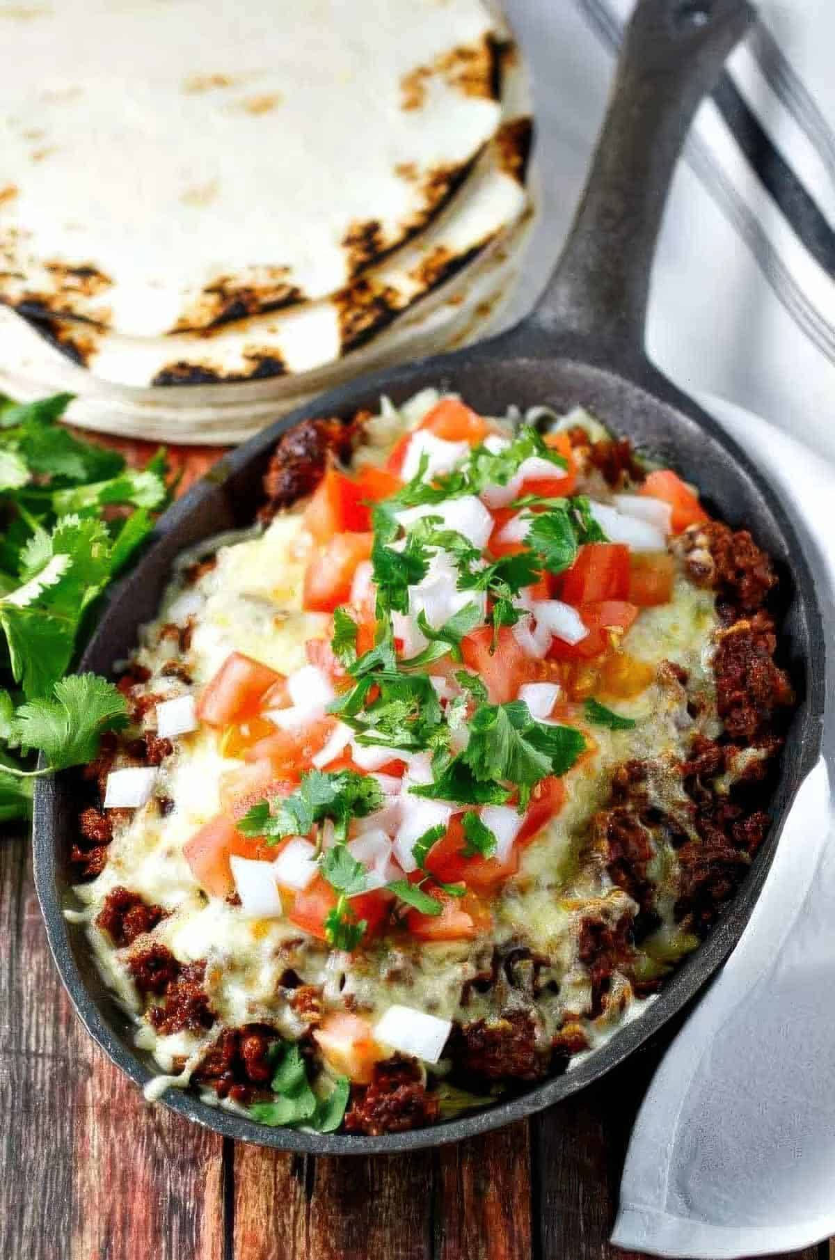 Choriqueso – Easy Mexican Chorizo and Cheese Dip a cast iron skillet filled with choriqueso made with chorizo and cheese then topped with diced tomato, white onion, and cilantro, next to a stack of charred flour tortillas