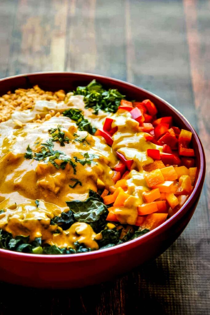 a very colorful chopped thai salad in a red bowl on a red table. the salad is covered in a coconut curry dressing with yellow and red bell peppers, kale, and chopped peanuts showing through the dressing