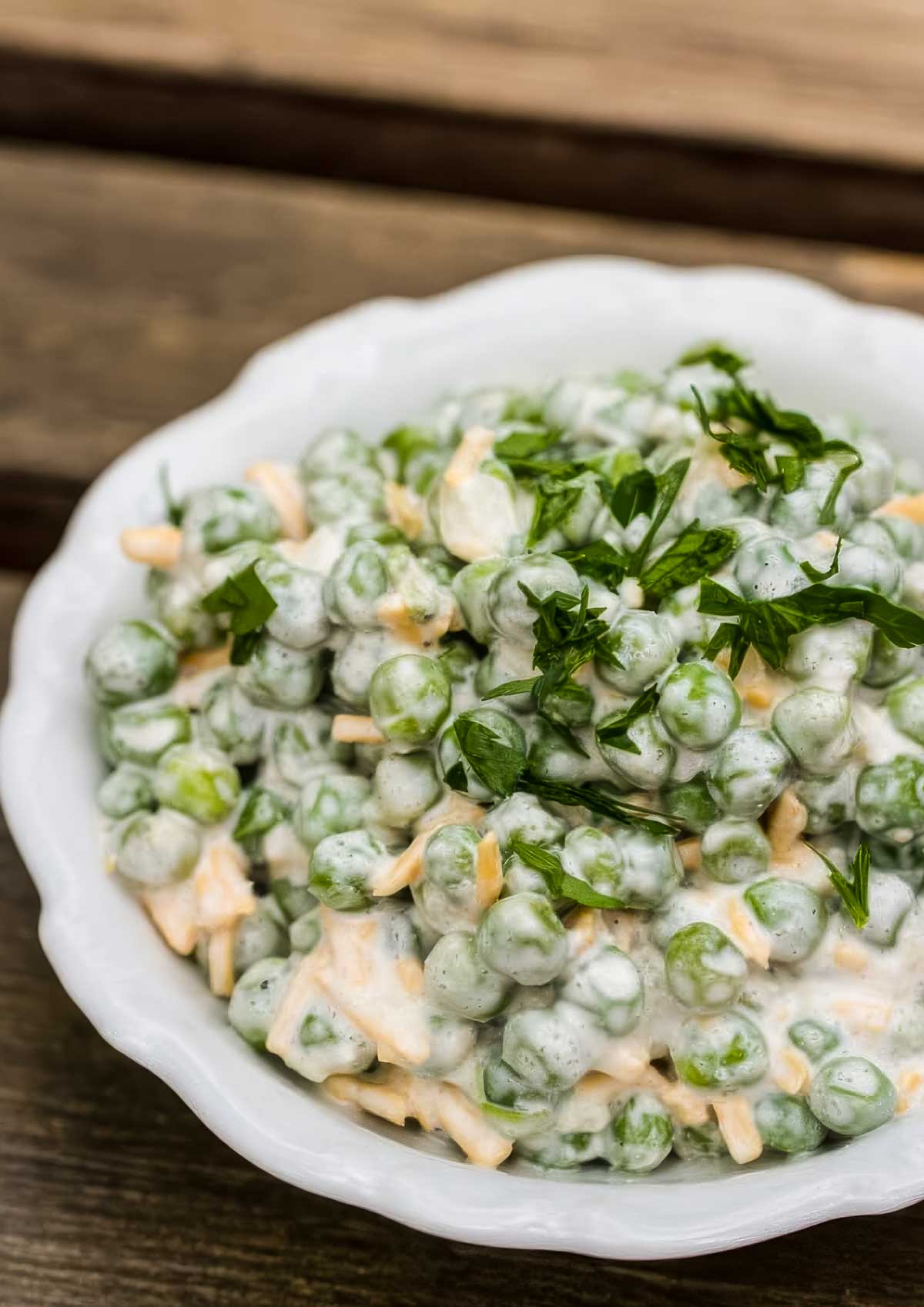 a white dish with scalloped edges, filled with an easy pea salad that's topped with chopped parsley on a wooden table