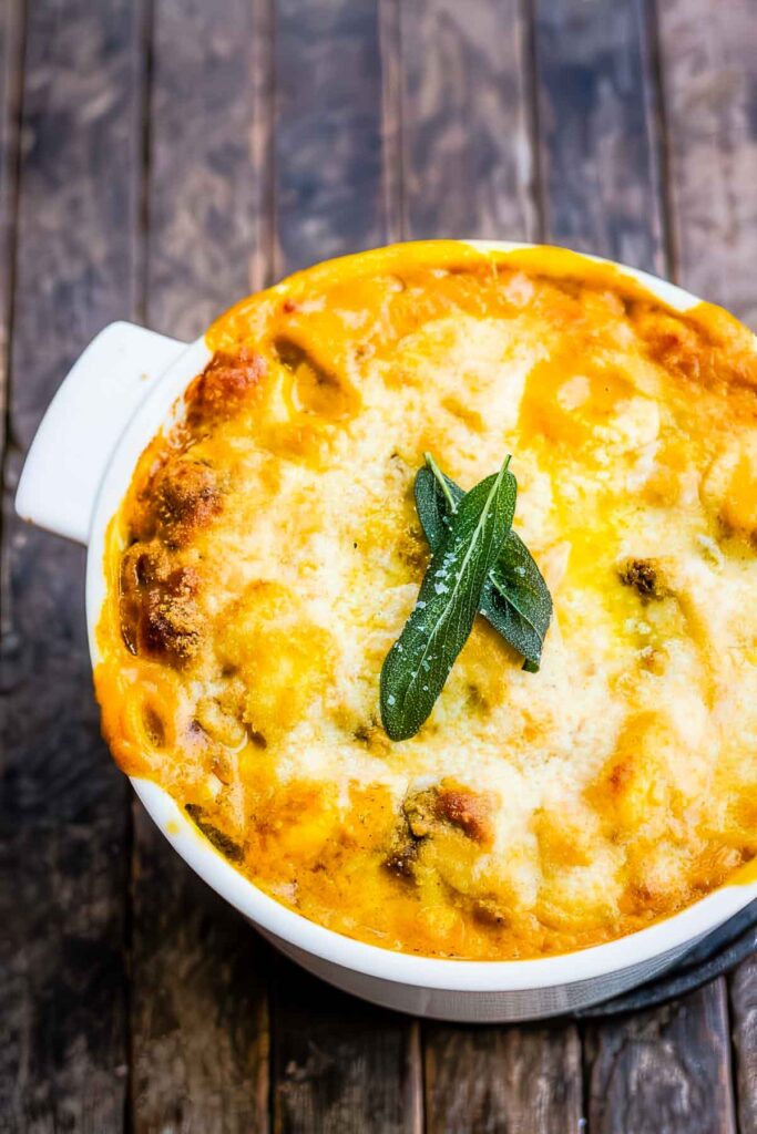 Golden baked pumpkin casserole topped with crispy sage leaves, presented in a white dish on a wooden table.