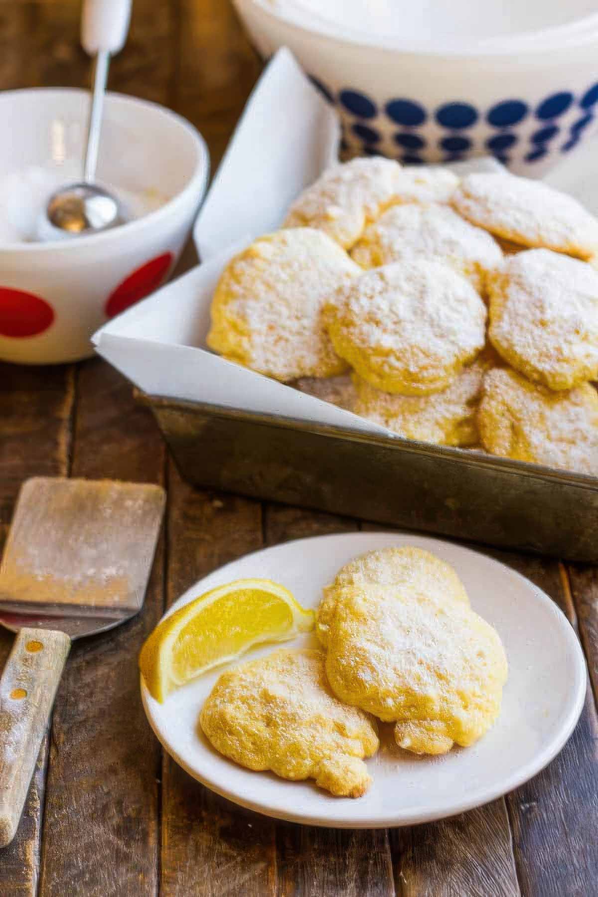 a small white plate of cake mix cookies with a fresh lemon wedge next to a vintage metal baking pan lined with parchment paper and filled with lemon cake mix cookies sprinkled with powdered sugar
