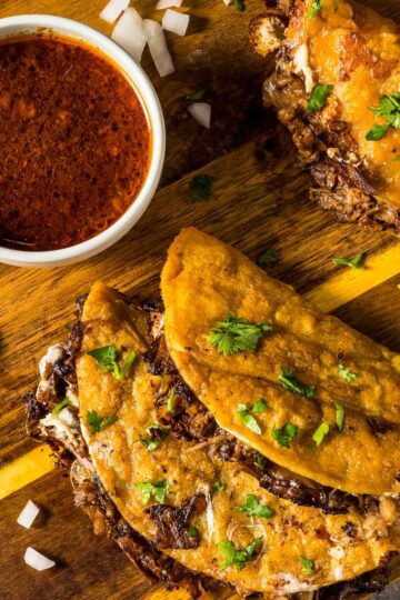 top down view of two birria tacos on a wooden board next to a small bowl of consommé