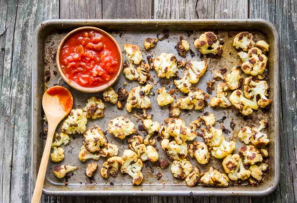 Roasted cauliflower served with a small bowl of marinara sauce and a wooden spoon on the pan.