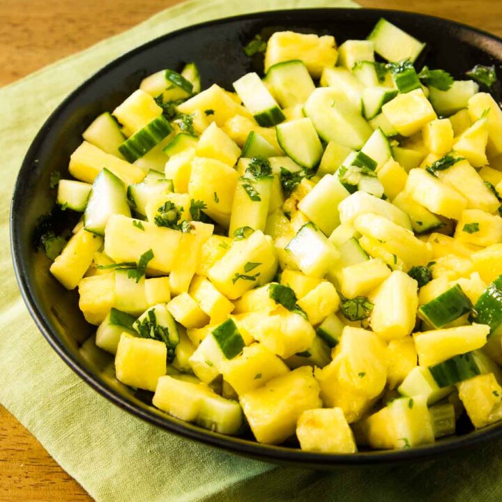 a black serving bowl filled with cucumber and pineapple salad with hot honey and cilantro, it's sitting on a green fabric napkin on a wooden table