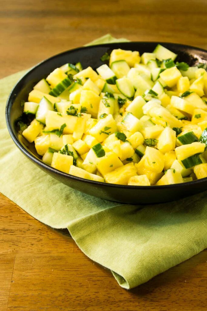 a black serving bowl filled with cucumber and pineapple salad with hot honey and cilantro, it's sitting on a green fabric napkin