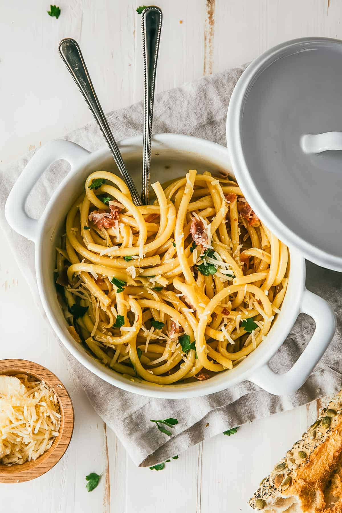 a closeup, overhead shot of an authentic Italian pasta carbonara recipe in a white ceramic pot with two serving spoons, on a white tablecloth with crusty bread
