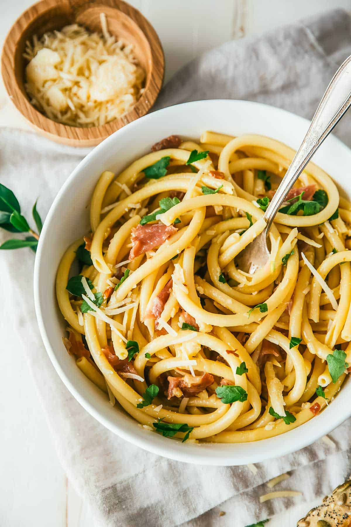 Authentic Italian Pasta Carbonara Recipe an overhead shot of an authentic Italian pasta carbonara recipe in a white bowl with a spoon, on a white tablecloth with crusty bread