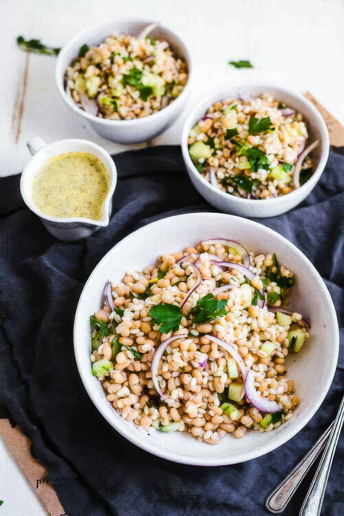 White Bean & Barley Salad with Lemon-Garlic Vinaigrette an overhead view of a white serving bowl filled with barley salad with white beans next to two smaller bowls on a black linen cloth next to a container of greek vinaigrette