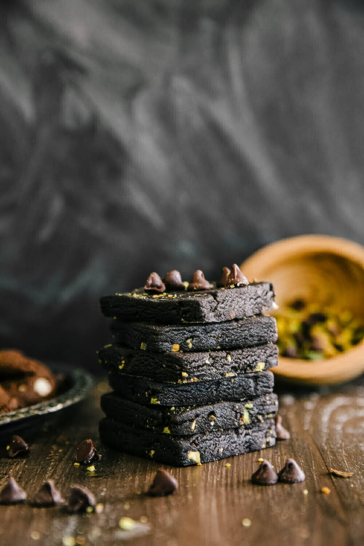 a stack of chocolate shortbread cookies with pistachios, with a few chocolate chips sprinkled on top and a small wooden bowl of pistachios in the background