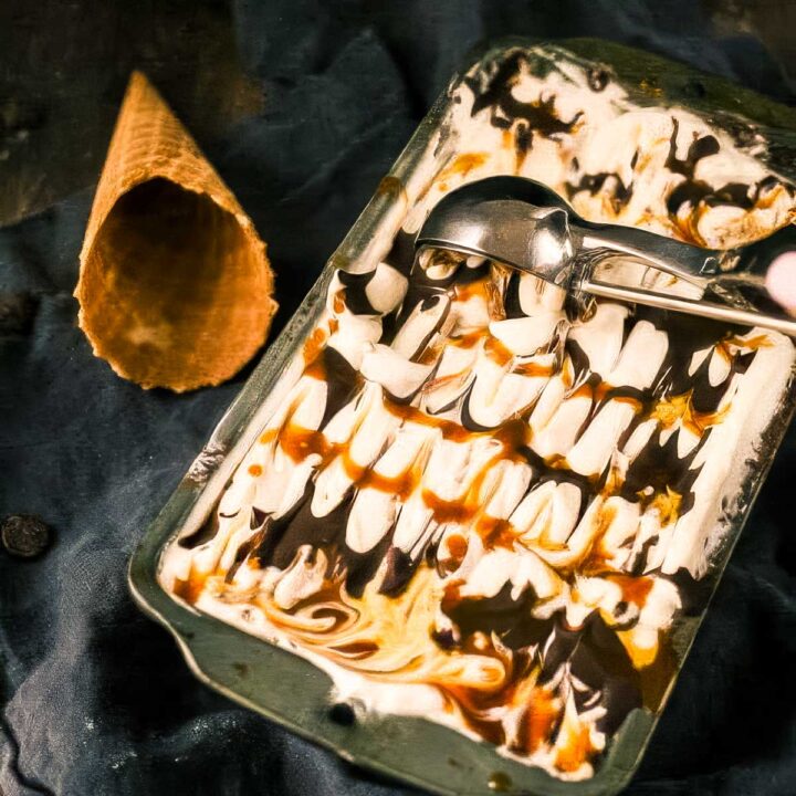 a metal loaf pan filled to the brim with chunky monkey ice cream, with an ice cream scoop sitting on the top and a waffle cone next to it