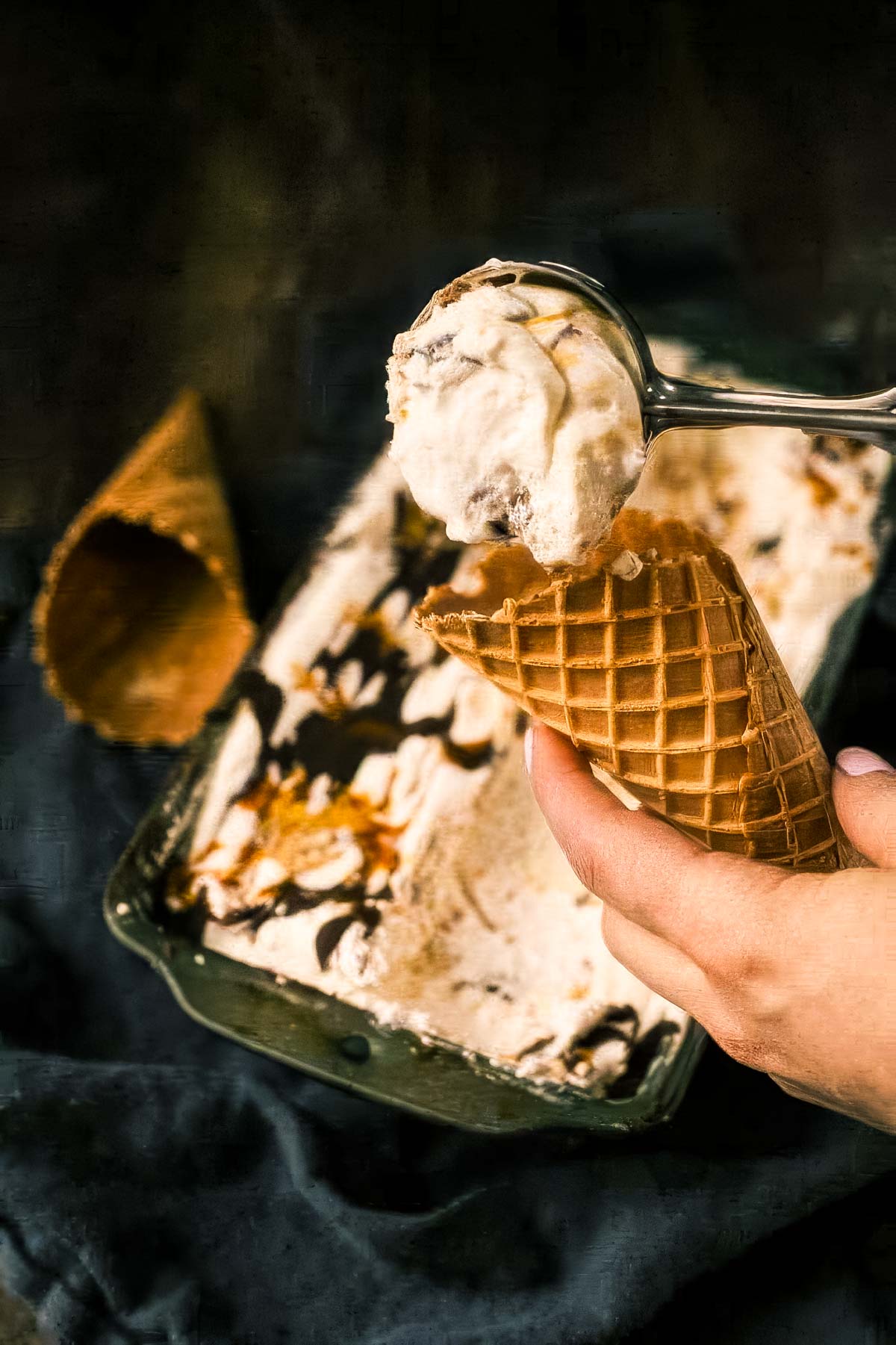 Chunky Monkey Ice Cream Recipe (No-Churn!) a hand holding a waffle cone with a scoop of chunky monkey ice cream being placed in it