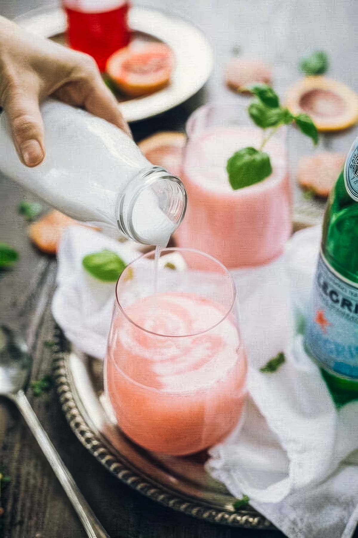 coconut milk being poured into a glass of italian blood red soda garnished with fresh basil, on a tray next to a bottle of pellegrino and fresh blood orange slices
