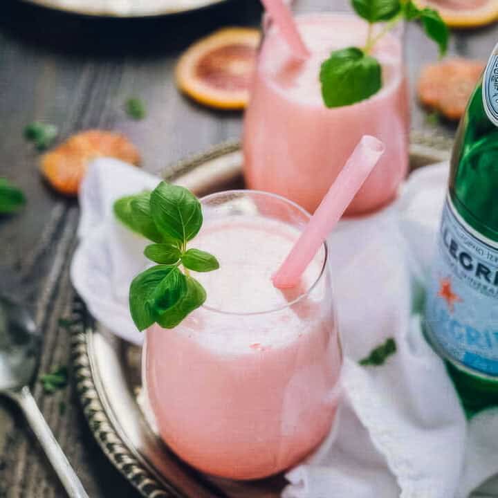 a closeup of two glasses of italian blood red soda garnished with fresh basil, on a tray next to a bottle of pellegrino and fresh blood orange slices